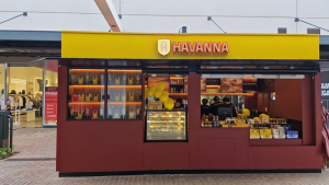 Quiosque da marca Havanna com fachada amarela e vermelha, exibindo produtos como doces e embalagens, decorado com balões amarelos, localizado em área comercial ao ar livre.