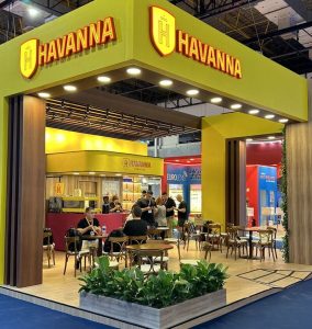 Estande da Havanna em evento, com estrutura moderna em tons de amarelo e madeira, mesas para atendimento e pessoas reunidas ao redor do balcão.