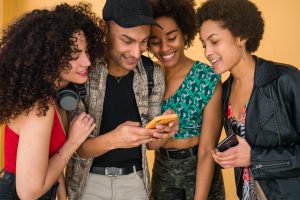 Grupo de quatro jovens amigos sorrindo e olhando para um celular, demonstrando empolgação e descontração.