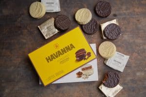 Caixa de alfajores Havanna aberta sobre uma mesa de madeira, cercada por alfajores de chocolate e chocolate branco, alguns ainda embalados.