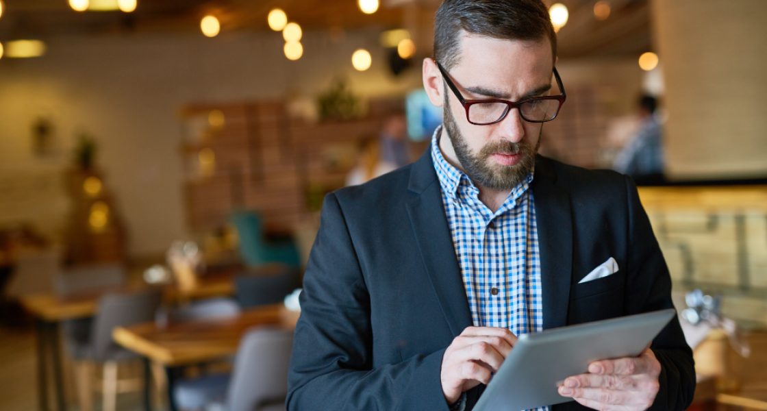 Homem com blazer preto e camisa xadrez usando um tablet em um ambiente moderno, com mesas e iluminação pendente ao fundo.