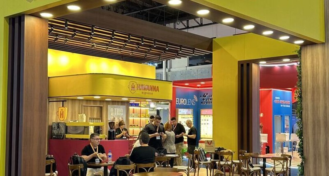 Estande da Havanna em evento, com estrutura moderna em tons de amarelo e madeira, mesas para atendimento e pessoas reunidas ao redor do balcão.