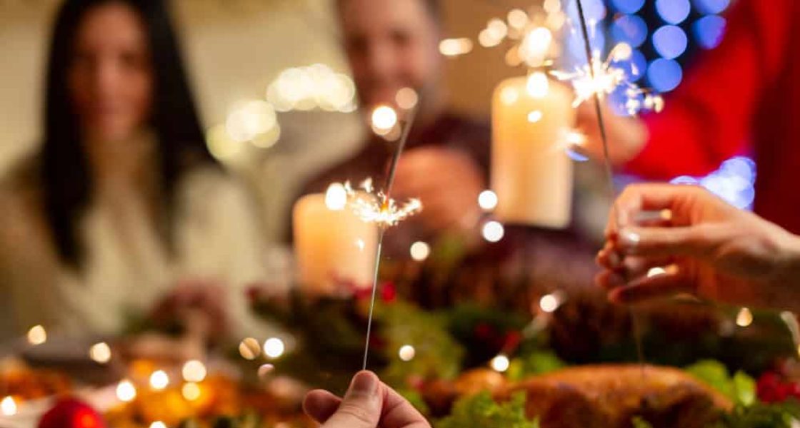 Pessoas celebrando a ceia de Natal com velas acesas, faíscas e pratos típicos em uma mesa decorada.