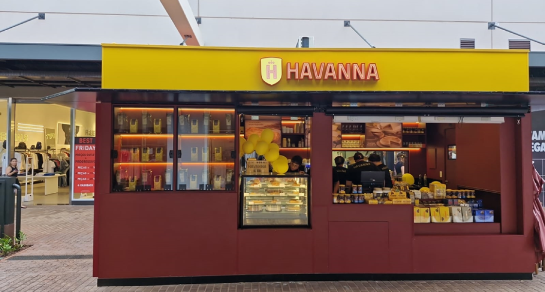 Quiosque da marca Havanna com fachada amarela e vermelha, exibindo produtos como doces e embalagens, decorado com balões amarelos, localizado em área comercial ao ar livre.