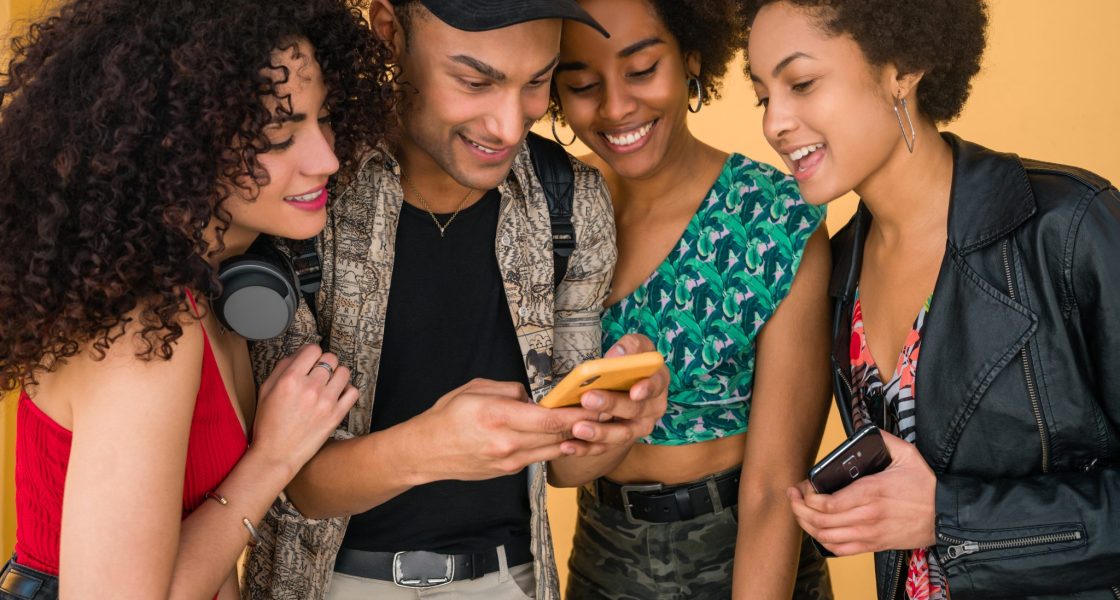 Grupo de quatro jovens amigos sorrindo e olhando para um celular, demonstrando empolgação e descontração.